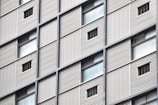 A close-up view of a building facade featuring a grid pattern with alternating solid panels and small window openings. The structure appears to have metal cladding with vertical lines providing a textured surface.