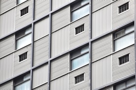 A close-up view of a building facade featuring a grid pattern with alternating solid panels and small window openings. The structure appears to have metal cladding with vertical lines providing a textured surface.