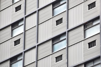 A close-up view of a building facade featuring a grid pattern with alternating solid panels and small window openings. The structure appears to have metal cladding with vertical lines providing a textured surface.
