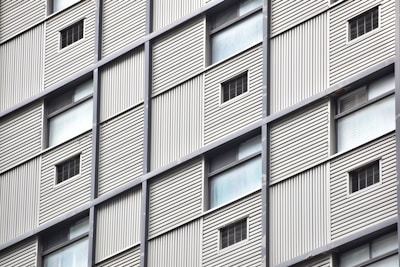 A close-up view of a building facade featuring a grid pattern with alternating solid panels and small window openings. The structure appears to have metal cladding with vertical lines providing a textured surface.