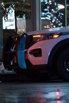 A police vehicle is parked on a wet urban street at night. The headlights are on, illuminating the area, and festive string lights are wrapped around a nearby tree, reflecting on the vehicle's exterior.