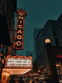 An illuminated marquee with the word 'Chicago' displayed vertically in bright lights, set against an urban cityscape with tall buildings. Below, performances including 'The Doobie Brothers' are listed. The scene is captured in the evening with visible streetlights and a dark teal sky.