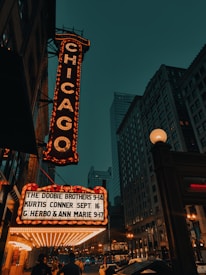 An illuminated marquee with the word 'Chicago' displayed vertically in bright lights, set against an urban cityscape with tall buildings. Below, performances including 'The Doobie Brothers' are listed. The scene is captured in the evening with visible streetlights and a dark teal sky.