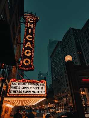 An illuminated marquee with the word 'Chicago' displayed vertically in bright lights, set against an urban cityscape with tall buildings. Below, performances including 'The Doobie Brothers' are listed. The scene is captured in the evening with visible streetlights and a dark teal sky.