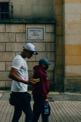Two people walking past a stone wall with a street sign that reads 'Primera Calle de la Carrera'. One person is wearing a white cap and T-shirt while looking at a phone, and the other person is wearing a maroon hoodie and mask, holding a water bottle.