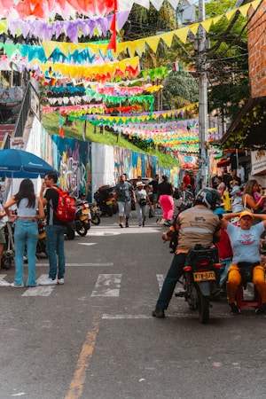 A lively urban street scene with colorful triangular flags hanging overhead. Graffiti covers the wall on the left side, and a bustling crowd of people, some on motorcycles, fill the street. Vendors are visible along the roadside, and the atmosphere is vibrant and festive.