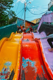 A group of children are sitting at the top of a colorful, multi-lane slide, appearing eager to slide down. The slide features vibrant colors such as yellow, red, and purple, with visible wear and scratches that add character. In the background, there are colorful buildings with conspicuous electrical wires overhead. Lush green trees frame part of the scene.