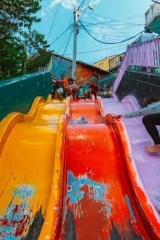 A group of children are sitting at the top of a colorful, multi-lane slide, appearing eager to slide down. The slide features vibrant colors such as yellow, red, and purple, with visible wear and scratches that add character. In the background, there are colorful buildings with conspicuous electrical wires overhead. Lush green trees frame part of the scene.