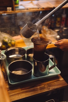 A person is pouring hot water from a metal ladle into a cloth filter placed over a kettle. The setup includes a tray with several metal containers, suggesting a coffee or tea preparation setting. Steam indicates the heat of the water, and the wooden counter adds warmth to the environment.
