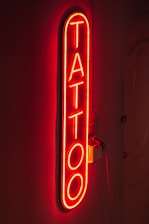 A bright red neon sign displaying the word 'TATTOO' vertically on a dark background, creating a glowing effect that highlights the word against the wall.