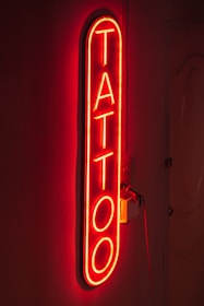 A bright red neon sign displaying the word 'TATTOO' vertically on a dark background, creating a glowing effect that highlights the word against the wall.