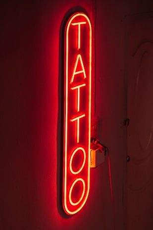 A bright red neon sign displaying the word 'TATTOO' vertically on a dark background, creating a glowing effect that highlights the word against the wall.