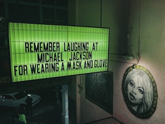 A green illuminated sign displays the text 'REMEMBER LAUGHING AT MICHAEL JACKSON FOR WEARING A MASK AND GLOVE'. Nearby, there is a black and white portrait of a woman on the wall, and below, people and a white car are visible in a dimly lit area.