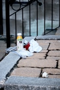 A small pile of items is placed on a stone ledge, including a bottle of orange juice, a red can, white cloths, and a plastic bag containing small objects. The background features a metal railing and a concrete wall, giving an urban feel to the setting.