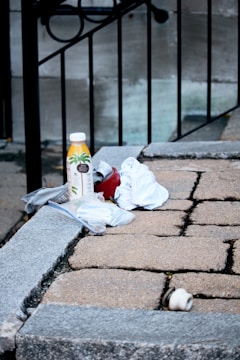 A small pile of items is placed on a stone ledge, including a bottle of orange juice, a red can, white cloths, and a plastic bag containing small objects. The background features a metal railing and a concrete wall, giving an urban feel to the setting.