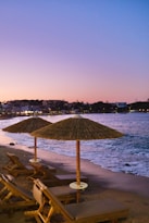 Sunset view of a serene beach with wooden lounge chairs and straw umbrellas. The shoreline curves gently with waves lapping at the sand. In the background, a town is visible, illuminated by lights as dusk falls. The sky transitions from pink to deep blue, creating a tranquil and picturesque scene.