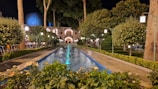 A beautifully landscaped garden at night, featuring a long rectangular water fountain with blue lighting. The pathway is lined with neatly trimmed bushes and trees, and ornate street lamps cast a soft glow over the scene. In the background, a large architectural structure with arches is partially visible, adding an element of grandeur.