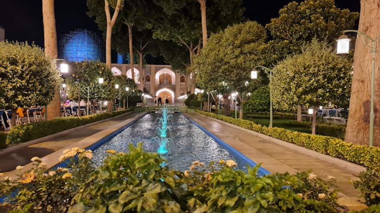 A beautifully landscaped garden at night, featuring a long rectangular water fountain with blue lighting. The pathway is lined with neatly trimmed bushes and trees, and ornate street lamps cast a soft glow over the scene. In the background, a large architectural structure with arches is partially visible, adding an element of grandeur.