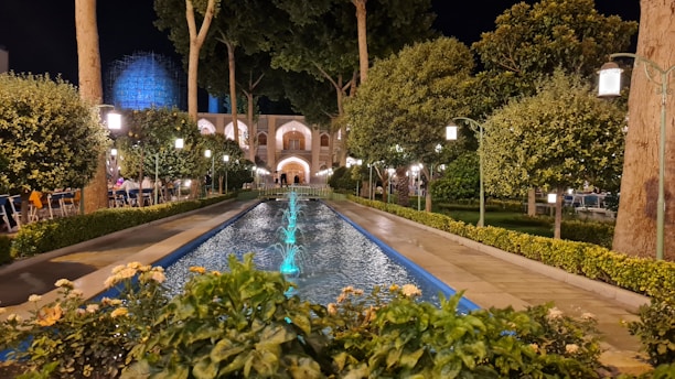 A beautifully landscaped garden at night, featuring a long rectangular water fountain with blue lighting. The pathway is lined with neatly trimmed bushes and trees, and ornate street lamps cast a soft glow over the scene. In the background, a large architectural structure with arches is partially visible, adding an element of grandeur.