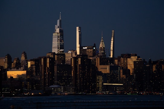 A city skyline at dusk featuring a variety of illuminated skyscrapers. The sky appears to be getting dark as night approaches, but some buildings reflect the warm glow of the setting sun. The image captures a blend of modern and older architectural designs with prominent high-rises.