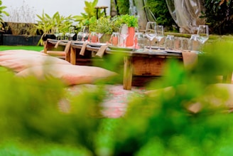 A wooden outdoor dining table is set on a vibrant green lawn, surrounded by lush plant life. The table is elegantly dressed with glassware, including wine glasses and water glasses, along with vases holding greenery. Large, soft cushions are placed on the ground for seating, providing a relaxed and cozy atmosphere.