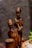 Wooden sculptures depict two figures with elongated features and a polished dark surface. One figure is seated, holding a stick, while the other stands nearby. They appear to be carved from a single piece of wood.