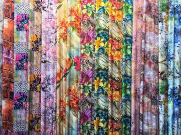 A vibrant collection of fabric rolls displayed vertically, featuring a variety of patterns and colors. The patterns include floral designs, abstract motifs, and geometric elements, with an array of colors such as bright yellow, pink, blue, and green.