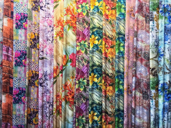 A vibrant collection of fabric rolls displayed vertically, featuring a variety of patterns and colors. The patterns include floral designs, abstract motifs, and geometric elements, with an array of colors such as bright yellow, pink, blue, and green.