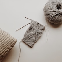 A knitted piece with a detailed cable pattern, accompanied by knitting needles and a ball of grey yarn. The setup is placed on a light surface with a patterned fabric cushion nearby.