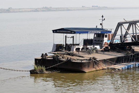 A large barge is moored on calm waters. It has a blue canopy with the name 'M.V. Kautilya Vihar' printed on it. Various ropes and chains are used to secure the vessel, and there are several chairs and a table on its deck. A control cabin is visible, and the surrounding water is wide and placid with a distant shoreline.