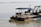 A large barge is moored on calm waters. It has a blue canopy with the name 'M.V. Kautilya Vihar' printed on it. Various ropes and chains are used to secure the vessel, and there are several chairs and a table on its deck. A control cabin is visible, and the surrounding water is wide and placid with a distant shoreline.