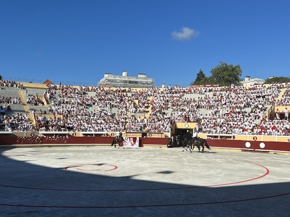 A crowded bullring with spectators dressed in white and red sitting on stadium-style seats, under a clear blue sky. Two mounted individuals can be seen in the arena, preparing for an event.