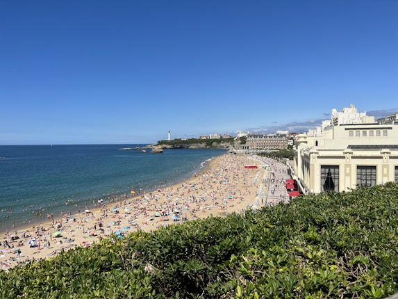 A sandy beach filled with numerous people relaxing under colorful umbrellas, adjacent to a large body of blue water. A lighthouse stands in the distance, and a clear, sunny sky stretches above. A grand, light-colored building borders the beach on the right side.