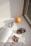 Three scrunchies of different colors are placed on a white surface next to an orange. An open book with text and images is also in view, positioned near a window with sunlight streaming in.