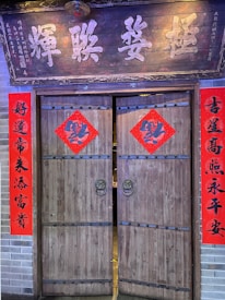 A wooden traditional Chinese door is adorned with red banners and decorations featuring Chinese calligraphy. The top features an ornate wooden sign with large, painted Chinese characters. Red diamond-shaped papers with additional characters are placed on each door panel. The surrounding wall is made of gray brick.