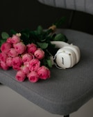 A bouquet of vibrant pink roses lies on a textured gray chair. Beside the flowers, there is a pair of white headphones with gold accents, adding a modern touch to the soft and romantic setting.