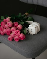A bouquet of vibrant pink roses lies on a textured gray chair. Beside the flowers, there is a pair of white headphones with gold accents, adding a modern touch to the soft and romantic setting.