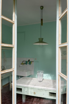 A kitchen space with mint green walls is seen through a partially open window. The room features a simple white sink with cleaning tools such as gloves and a sponge placed on the tiles. A marble countertop holds a large, decorative glass vase. An overhead light fixture with a retro design hangs from the ceiling. The overall atmosphere is calm and minimalistic.