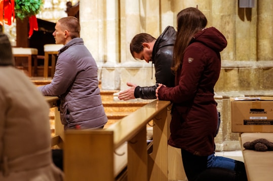 Three people are kneeling in prayer in a church. They are wearing winter jackets, and appear to be in a moment of reflection or meditation. The setting has wooden pews and stone walls, with a wreath and red ribbons in the background, suggesting a holiday season.