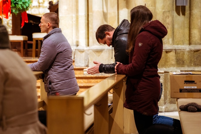 Three people are kneeling in prayer in a church. They are wearing winter jackets, and appear to be in a moment of reflection or meditation. The setting has wooden pews and stone walls, with a wreath and red ribbons in the background, suggesting a holiday season.