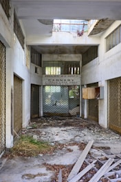 An abandoned and dilapidated interior of a shop with debris scattered on the floor. The shop has a metal gate, and there is a sign that reads 'Pavlos Pierides - Carnaby - Cyprus Lace Shop - Handicrafts & Souvenirs.' The walls and ceiling show signs of damage and decay, with exposed concrete and peeling paint. Natural light enters through broken windows, and there is overgrown grass and plants among the debris.