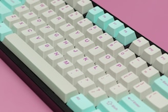 A mechanical keyboard with pastel-colored keycaps arranged on a pink background. The keys are labeled with purple alphanumeric characters and symbols. The keyboard includes a mix of standard white and mint-cyan keys, creating a visually appealing contrast.