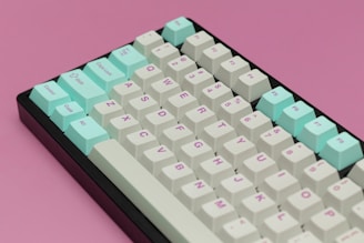 A mechanical keyboard with white and mint green keys is positioned on a pastel pink surface. The keys feature a mix of symbols and letters, and the layout appears to be standard QWERTY. The contrast between the colorful keys and the pink background creates a visually appealing aesthetic.
