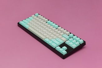A mechanical keyboard with a combination of teal and white keys arranged on a black frame is placed against a pink background. The keyboard shows a compact layout, and the different colored keys create a visually striking appearance.