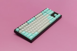 A mechanical keyboard placed on a solid pink background. The keyboard features a black base with pastel mint green and white keys. The keys are arranged in a compact layout, with function keys, letters, and numbers visible.
