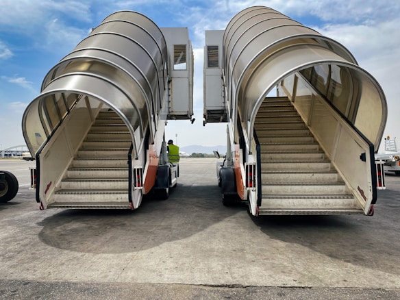 Two airport passenger boarding stairs with curved covers are parked on the tarmac, positioned side by side. A person in a high-visibility vest is standing near the stairs, against a backdrop of a partly cloudy sky and distant mountains.
