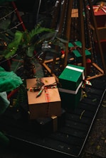 A festive arrangement of wrapped gift boxes with colorful ribbons is placed on a wooden platform. The scene is adorned with a small palm plant and set against a dark background, enhancing the vibrant colors of the gifts.
