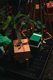A festive arrangement of wrapped gift boxes with colorful ribbons is placed on a wooden platform. The scene is adorned with a small palm plant and set against a dark background, enhancing the vibrant colors of the gifts.