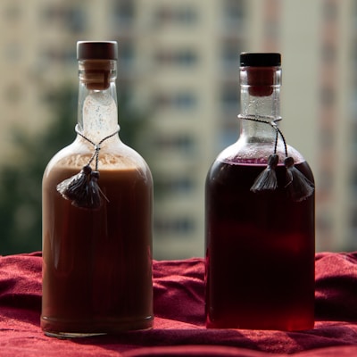 Two glass bottles are placed on a red fabric, with a beige-colored liquid in one and a dark red liquid in the other. Both bottles are sealed with corded stoppers, and the background features an out-of-focus urban skyline.