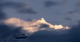 A snow-covered mountain peak rises prominently through a blanket of soft clouds. Warm sunlight illuminates the mountaintop, creating a striking contrast against the cooler, darker hues of the surrounding sky and clouds.